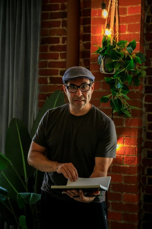 a man standing in front of a brick wall holding a book, tony khan wearing a headset, standing in a restaurant, next to a plant, profile image