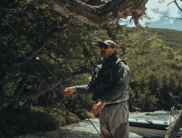 a man standing next to a boat on a river, patagonian, hunters gear, grey, wearing sunglasses and a cap