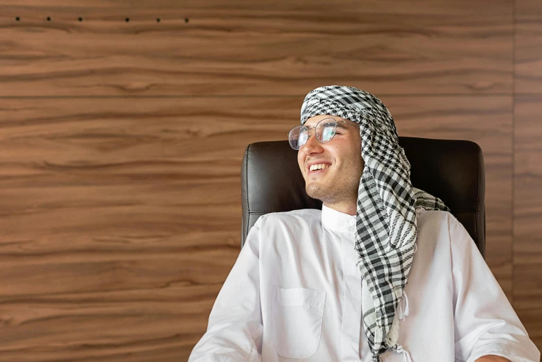 a man sitting in a chair with a scarf over his head, pexels contest winner, hurufiyya, corporate boss, wearing small round glasses, arab inspired, sarcastic smiling