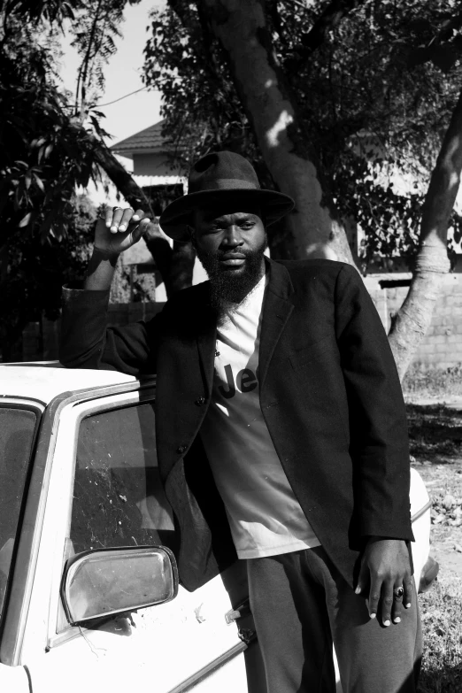 a black and white photo of a man leaning against a car, inspired by Mustafa Rakim, black beard, rasta, pulp fiction style, in a suburb