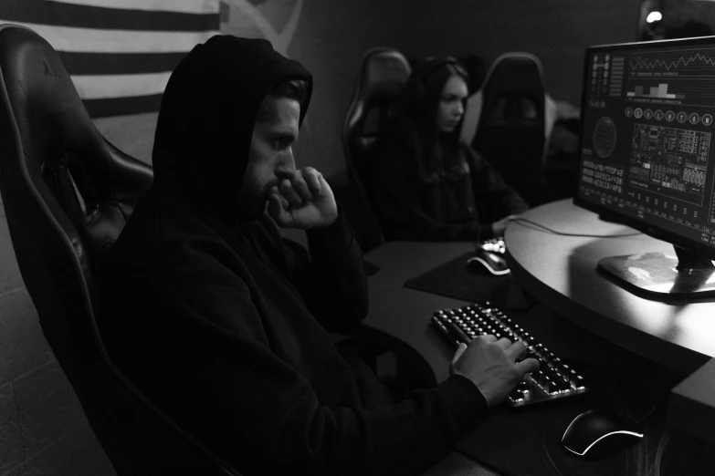 a man sitting at a desk in front of a computer, a black and white photo, by Adam Marczyński, game footage, squad, wearing a dark hood, they are very serious