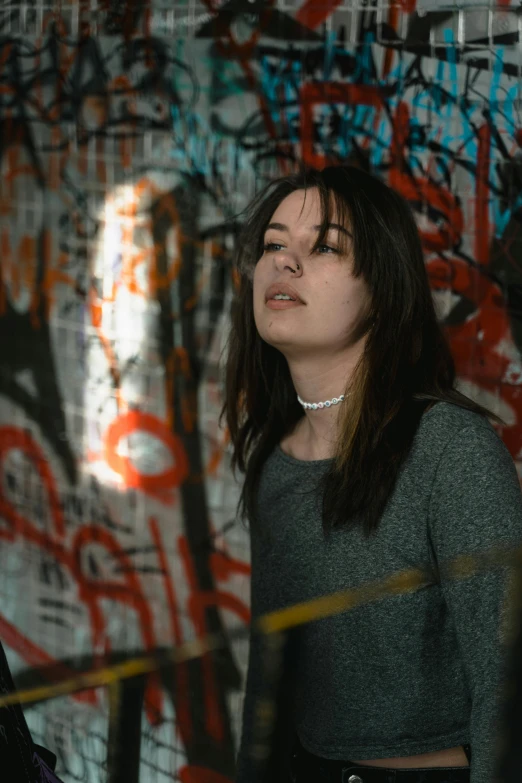 a woman standing in front of a graffiti covered wall, an album cover, inspired by Elsa Bleda, pexels contest winner, graffiti, curtain bangs, looking at the ceiling, portrait of depressed teen, concert photo