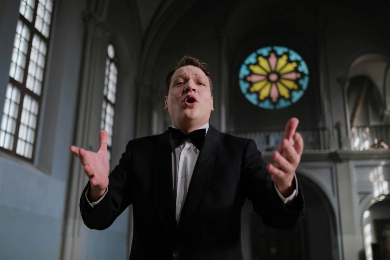 a man in a tuxedo standing in front of a stained glass window, an album cover, inspired by Emil Bisttram, pexels, baroque, singing at a opera house, in a church. medium shot, andy richter, kris kuksi