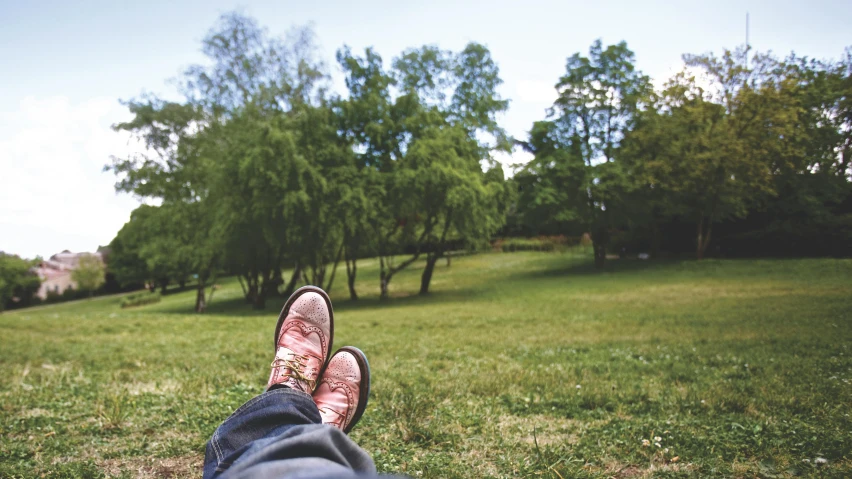 a person sitting on top of a lush green field, feet on the ground, in a park, relaxing on a couch, trees in background