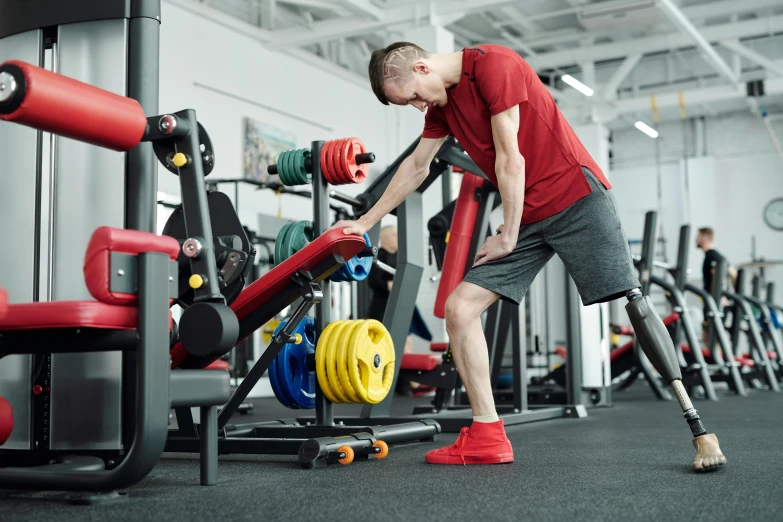 a man standing next to a machine in a gym, by Adam Marczyński, hurufiyya, two crutches near bench, high-quality photo, artificial limbs, train with maroon