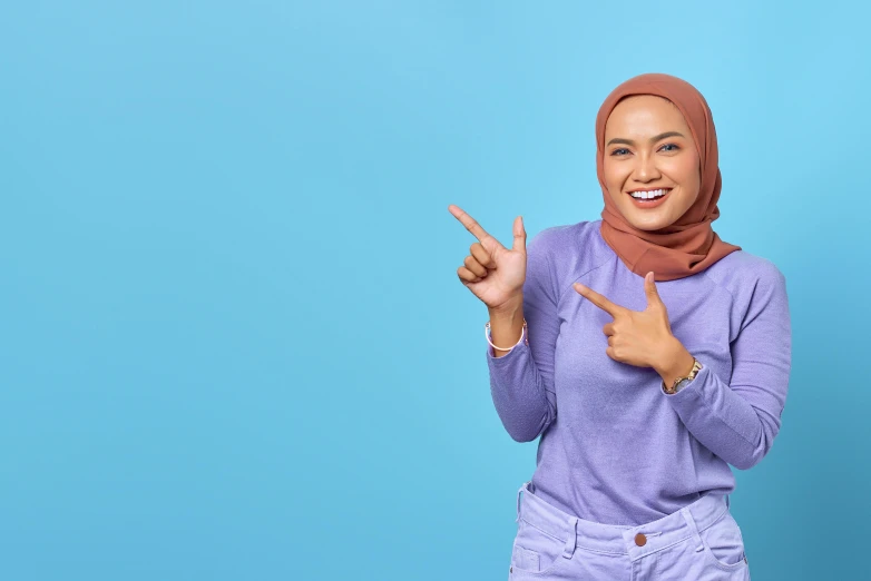 a woman in a hijab making a peace sign with her hands, a poster, inspired by JoWOnder, trending on pexels, hurufiyya, blue backdrop, pointing index finger, background image, marketing photo