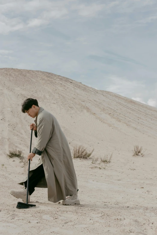 a man standing in the sand with a shovel, an album cover, inspired by Amir Zand, unsplash, wearing a long coat, asian male, heartbreak, ignant