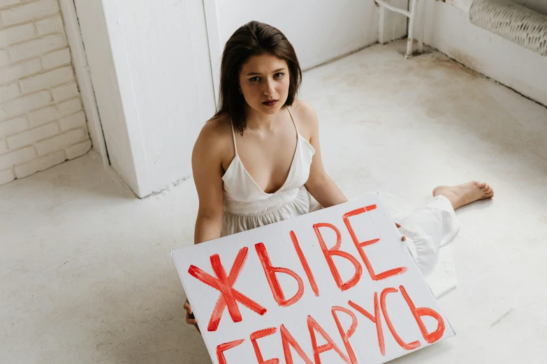 a woman sitting on the floor holding a sign, pexels contest winner, feminist art, russian, on a white table, 15081959 21121991 01012000 4k
