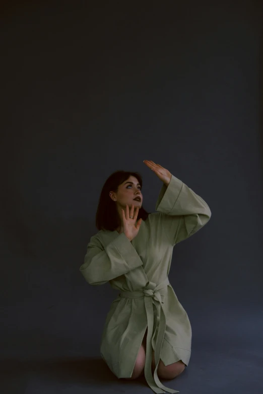 a woman in a green robe sitting on the ground, an album cover, by Attila Meszlenyi, unsplash, shrugging arms, grey backdrop, standing in a dimly lit room, wearing a track suit