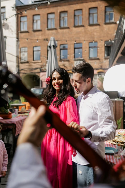 a man standing next to a woman in a pink dress, an album cover, by Julia Pishtar, pexels contest winner, happening, people outside eating meals, playing a mandolin, rooftop party, cheerful atmosphere