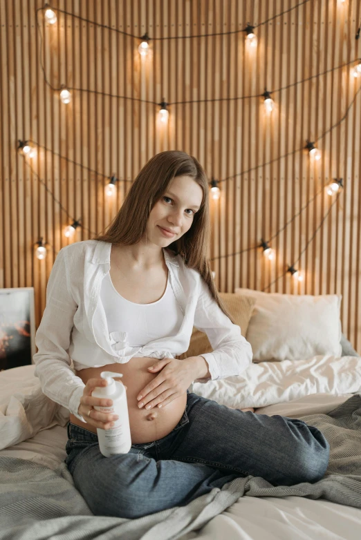 a woman sitting on top of a bed holding a cup, pregnant belly, multiple lights, skincare, backdrop