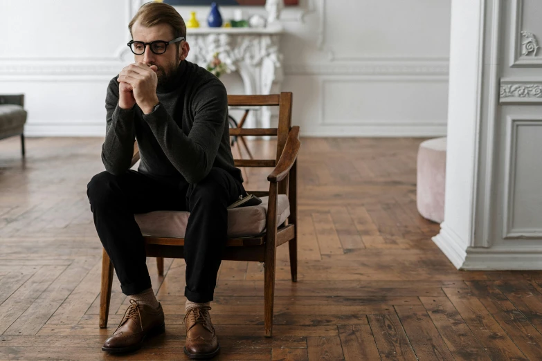 a man sitting on a chair in a room, inspired by Constantin Hansen, pexels, oiled hardwood floors, square rimmed glasses, alexandra fomina, thinking