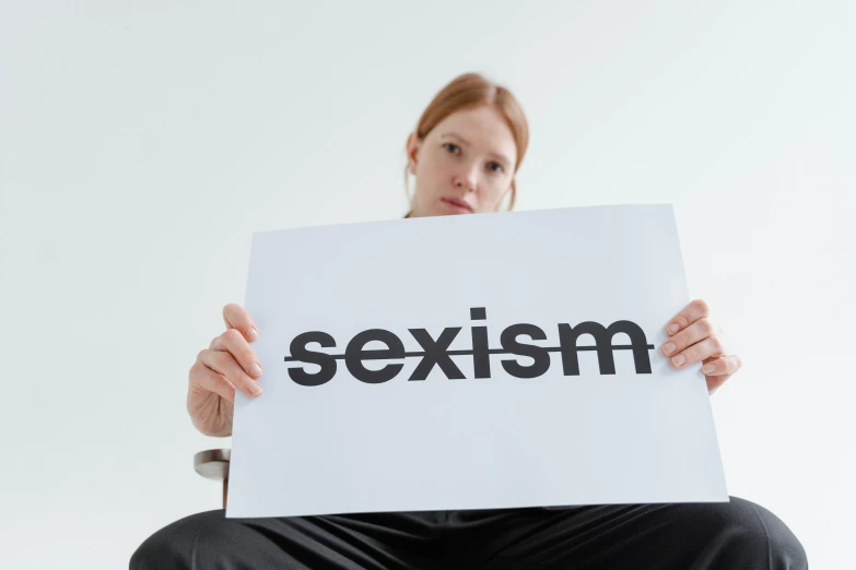 a woman holding a sign that says sexism, a poster, by Julia Pishtar, trending on pexels, on a pale background, studio photo, looking from slightly below, ecchi
