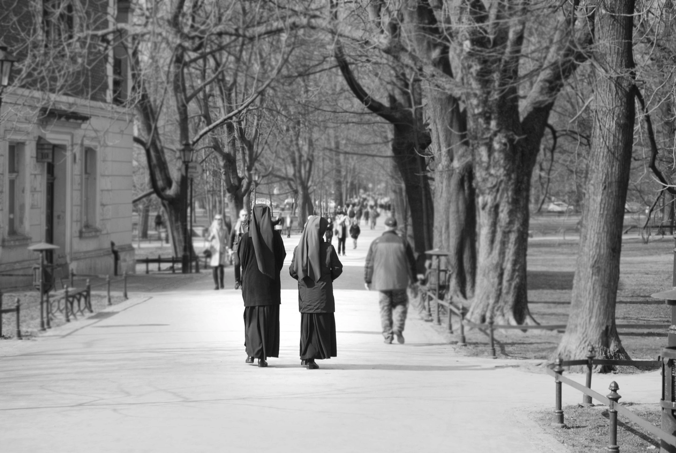 a black and white photo of two people walking down a sidewalk, two hovering twin nuns, berlin park, warm spring, romanian