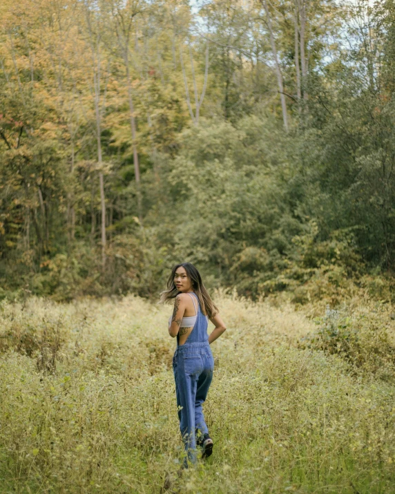 a woman standing in a field of tall grass, by Carey Morris, pexels contest winner, blue overalls, in serene forest setting, muted colored bodysuit, full body full height