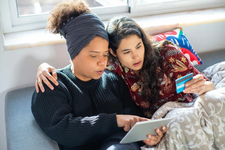 two women sitting on a couch looking at a tablet, trending on pexels, renaissance, aida muluneh, mourning family, bottom angle, holding each other