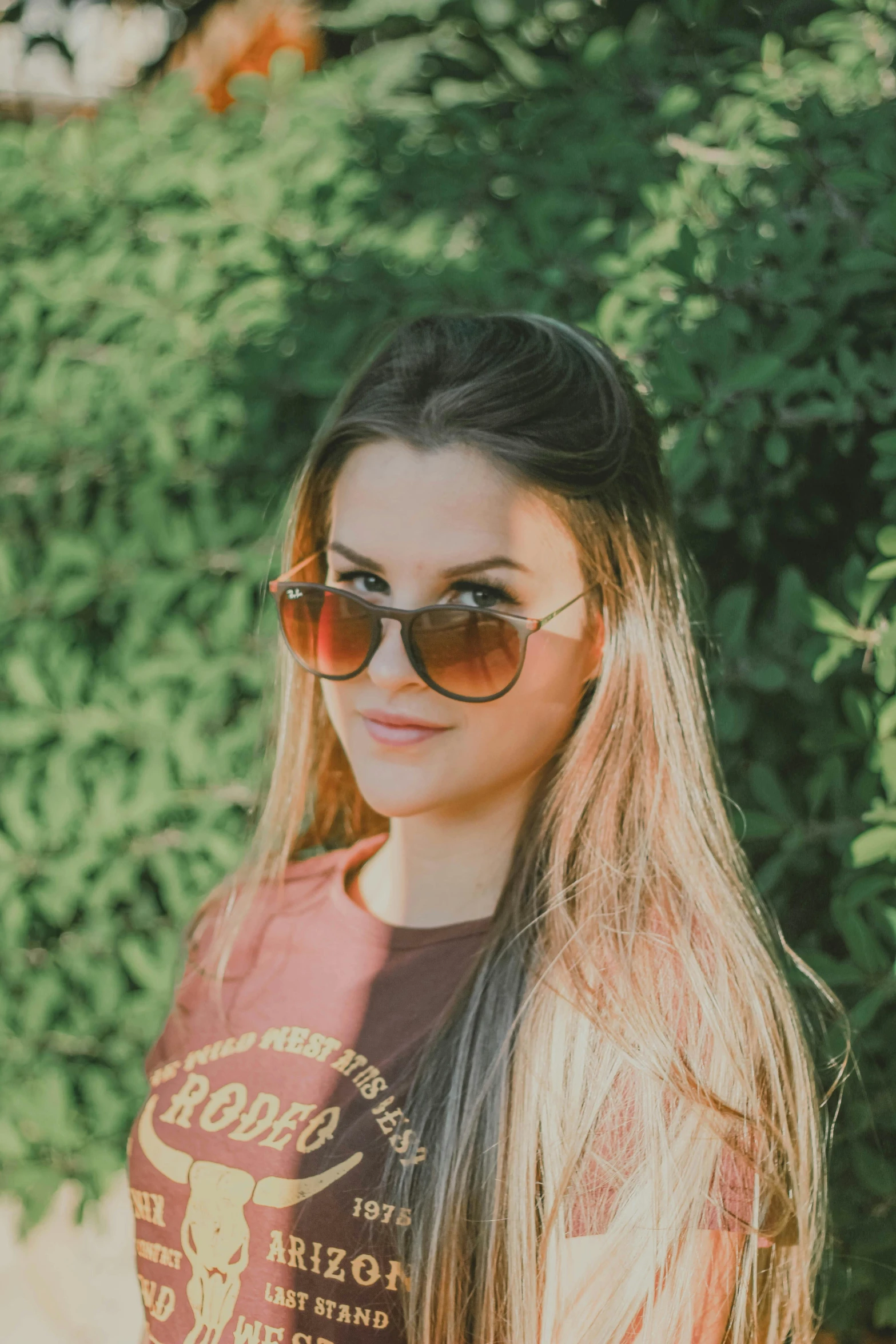 a woman wearing sunglasses standing in front of a bush, inspired by Elsa Bleda, unsplash, gradient brown to red, headshot profile picture, 18 years old, wear ray - ban glass