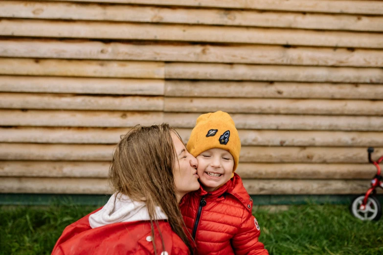 a woman sitting next to a little boy in a red jacket, a picture, by Julia Pishtar, pexels contest winner, yellow cap, stood outside a wooden cabin, marton gyula kiss ( kimagu ), full cheeks