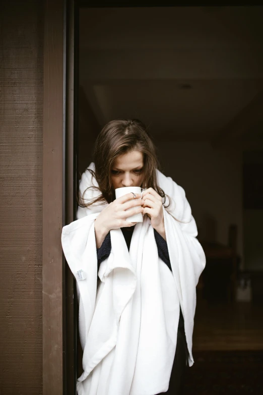 a woman standing in a doorway holding a cup of coffee, inspired by Elsa Bleda, pexels contest winner, happening, white robe, covered with blanket, woman very tired, girl with brown hair