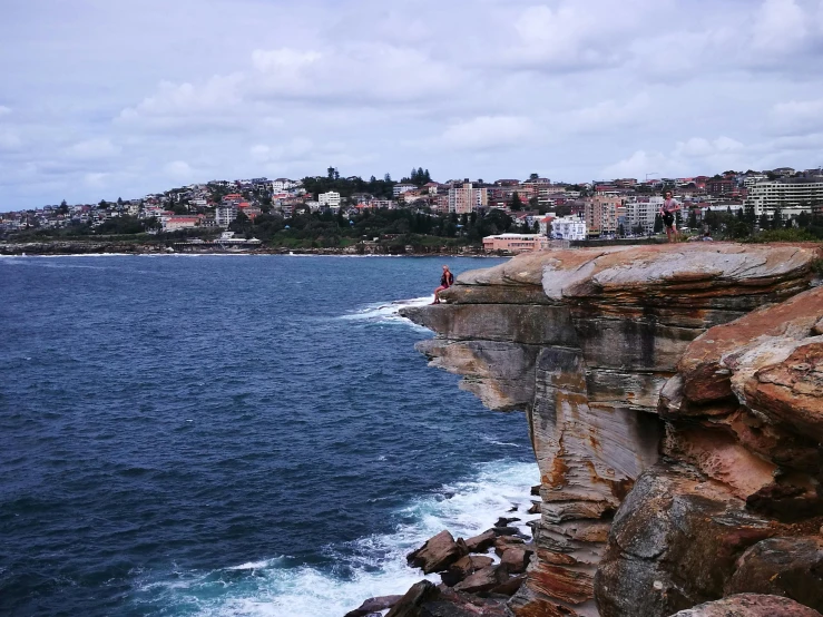 a man standing on top of a cliff next to the ocean, inspired by Sydney Carline, pexels contest winner, buildings carved out of stone, manly, geological strata, people walking around