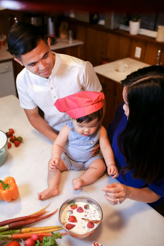 a man and woman in a kitchen with a baby, by Robbie Trevino, chef hat, mai anh tran, multicoloured, tabletop