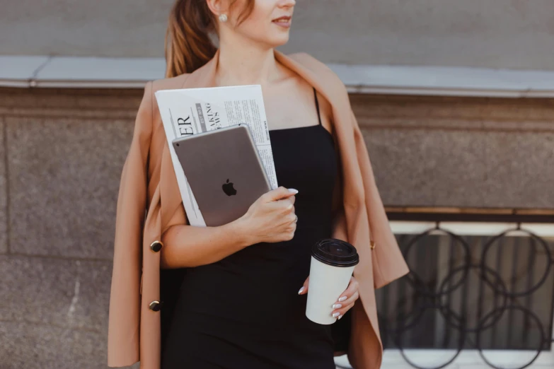 a woman holding a cup of coffee and a book, by Emma Andijewska, trending on pexels, private press, wearing black vest and skirt, newspaper, holding glowing laptop computer, light brown coat