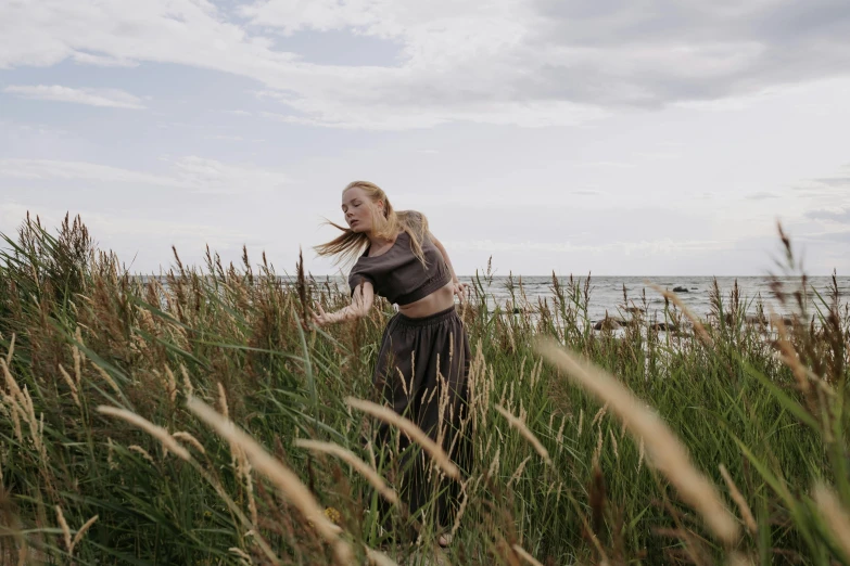 a woman standing in tall grass near a body of water, an album cover, by Emma Andijewska, unsplash, happening, dancing on the beach, greta thunberg, ignant, model posing