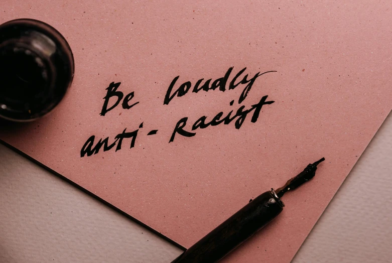 a pen sitting on top of a piece of paper, by Arabella Rankin, trending on pexels, antipodeans, black face, anti - communist, lettering, brown and pink color scheme