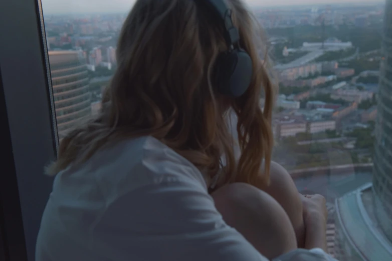 a woman wearing headphones looking out a window, inspired by Elsa Bleda, trending on unsplash, realism, sitting on a skyscraper rooftop, graded with davinci resolve, realistic », teaser