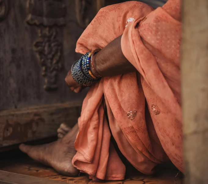 a close up of a person sitting on a wooden floor, inspired by Steve McCurry, trending on unsplash, dressed in a sari, clothed in ancient street wear, intricate african jewellery, bare leg