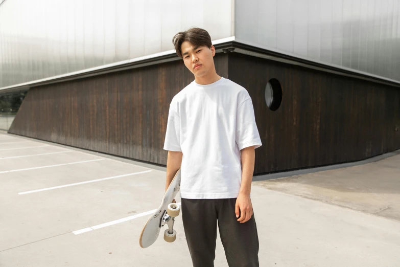 a young man holding a skateboard in a parking lot, inspired by Itō Ogura Yonesuke, unsplash, white t-shirt, in front of white back drop, wearing a baggy, detailed product image