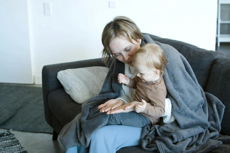 a woman sitting on a couch holding a baby, pexels, grey robe, covered with blanket, trying to read, gif