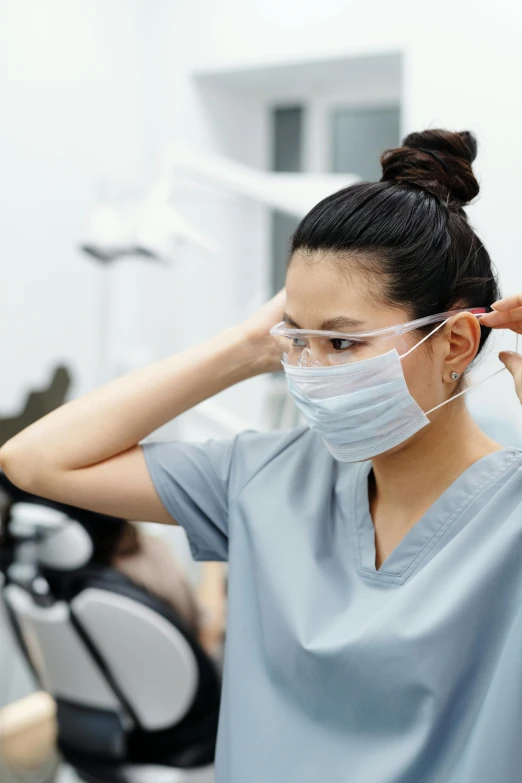 a woman wearing a face mask in a dentist's office, trending on pexels, happening, surgical gown and scrubs on, on grey background, round portruding chin, looking left