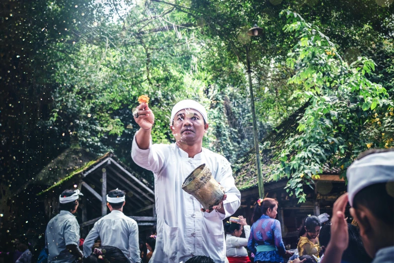 a man holding a bucket in front of a crowd of people, a portrait, inspired by I Ketut Soki, unsplash, sumatraism, ritual in a forest, 🕹️ 😎 🔫 🤖 🚬, the wonder of devotion, gong