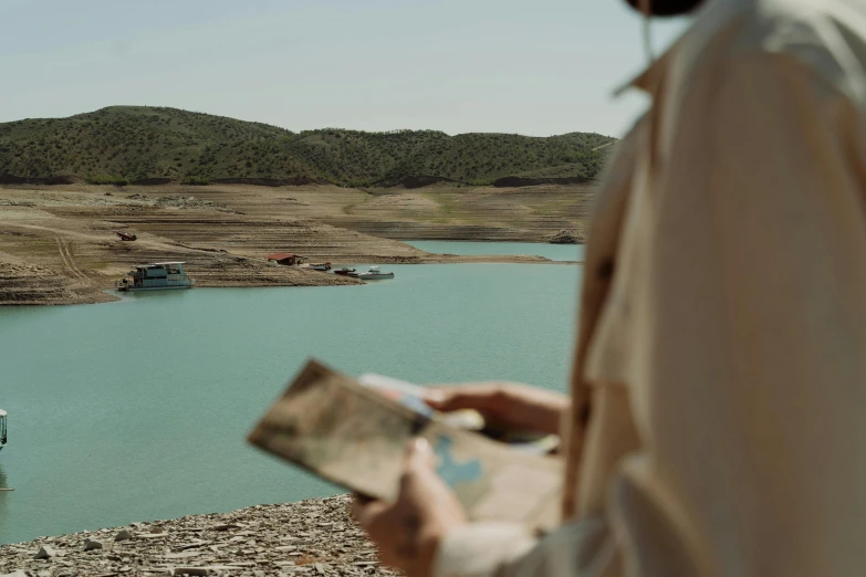 a man standing next to a lake holding a map, pexels contest winner, les nabis, oasis in the desert, cinestill colour cinematography, on a boat on a lake, teaser