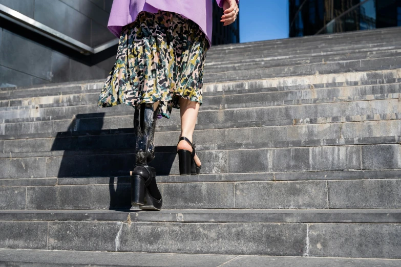 a woman walking up a flight of stairs, prosthetic, wearing floral chiton, wearing black boots, on a bright day