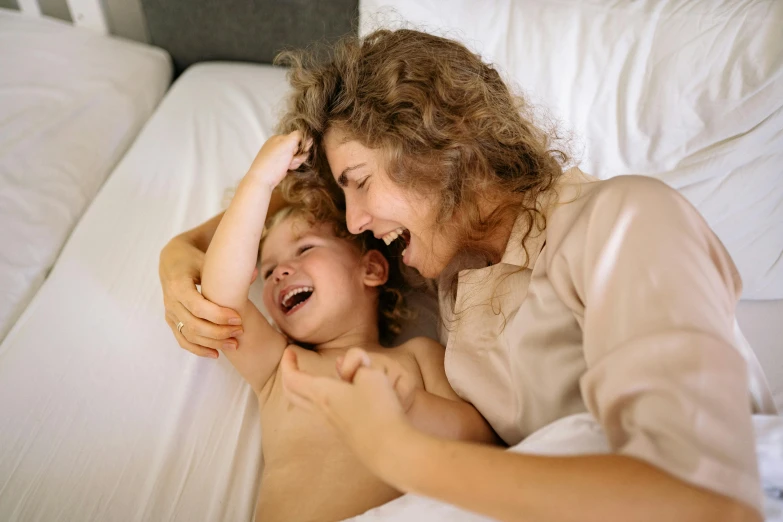 a woman laying on top of a bed next to a child, pexels contest winner, head bent back in laughter, 1 5 0 4, white, bedhead