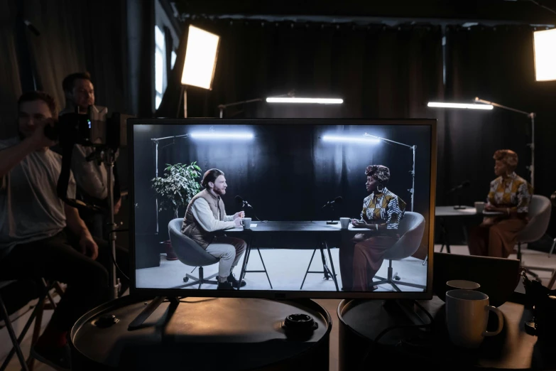 a group of people sitting around a table in front of a tv, a portrait, by Julia Pishtar, unsplash, video art, two still figures facing camera, in a dark studio room, set photo in costume, table with microphones