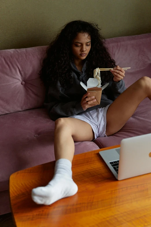 a woman sitting on a couch with a laptop, trending on pexels, renaissance, eating noodles, bra and shorts streetwear, thigh highs, in a taco bell