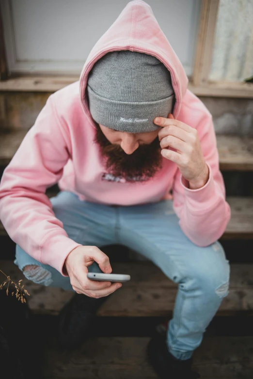 a man in a pink hoodie using a cell phone, by Adriaen Hanneman, trending on pexels, he also wears a grey beanie, denim, avatar image, sitting