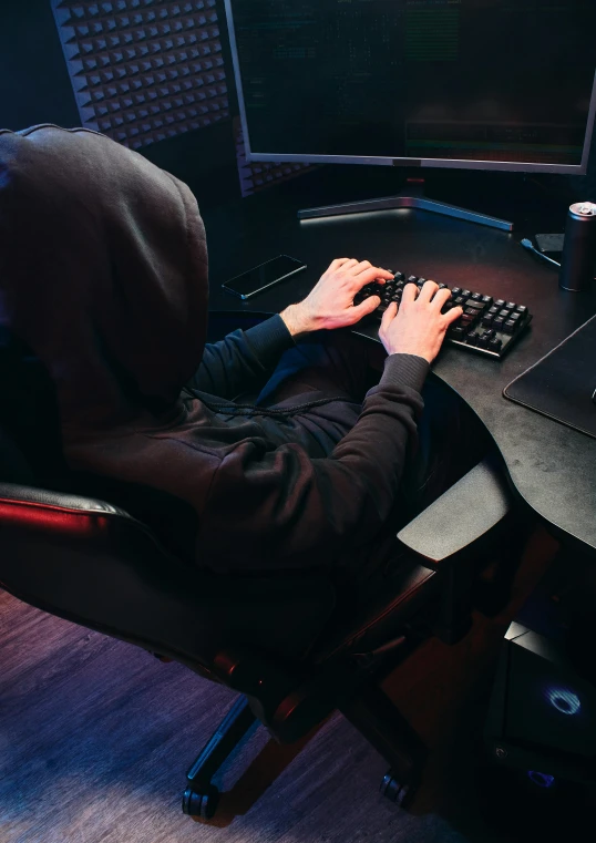 a person sitting at a desk in front of a computer, in a black hoodie, gaming chair, server in the middle, no - text no - logo
