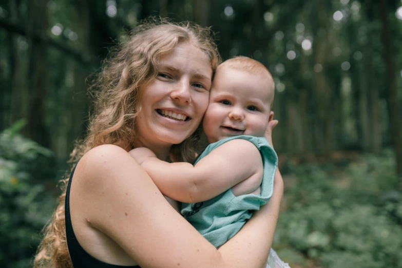 a woman holding a baby in a forest, colour portrait photograph, full frame image, avatar image, smiling