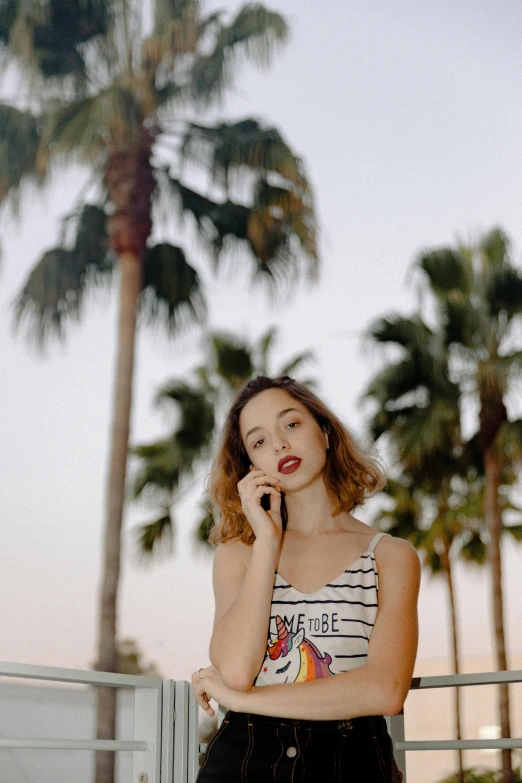 a woman talking on a cell phone next to palm trees, inspired by Elsa Bleda, trending on pexels, happening, kiko mizuhara, small and thick red lips, white! tank top with a print, striped