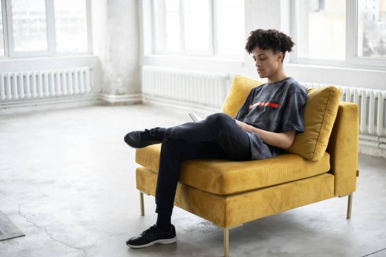 a young man sitting on top of a yellow couch, by Nina Hamnett, pexels contest winner, happening, graphic tees, nathalie emmanuel, reading, in a comfortable chair