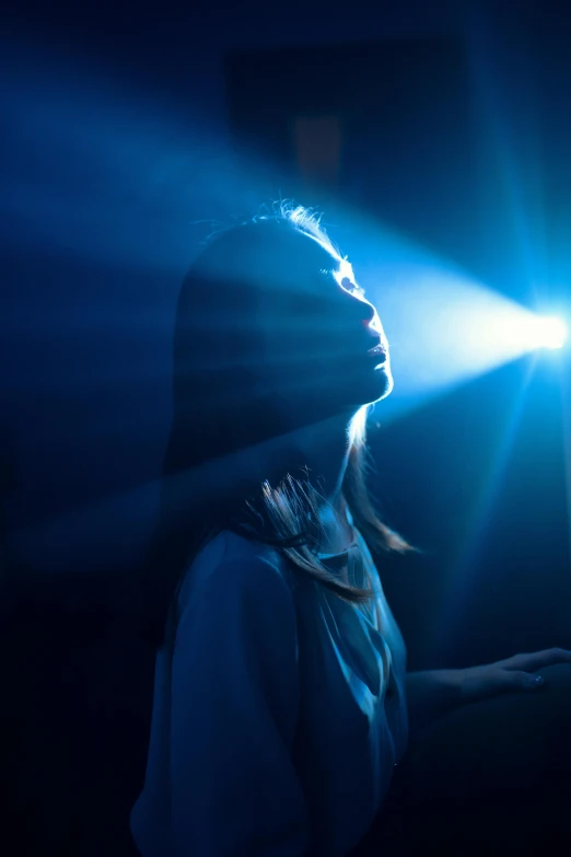 a woman standing in front of a bright light, a hologram, by David Donaldson, pexels contest winner, young woman looking up, dramatic blue light, holy rays, search lights