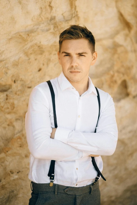 a man standing in front of a stone wall, trending on pexels, suspenders, zac efron, julian ope, wearing white shirt