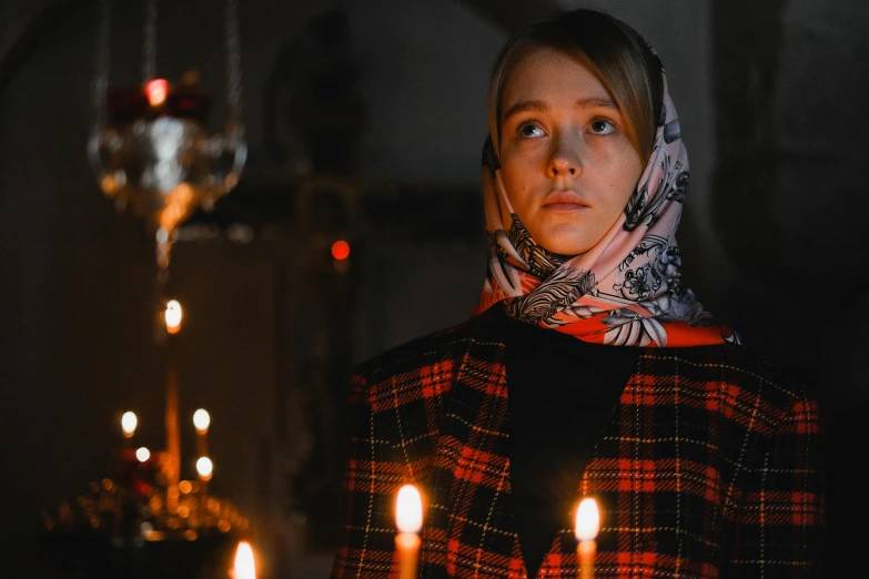 a woman standing in front of a bunch of lit candles, a portrait, by Julia Pishtar, pexels contest winner, hyperrealism, patterned scarf, scene from church, sadie sink, russian clothes