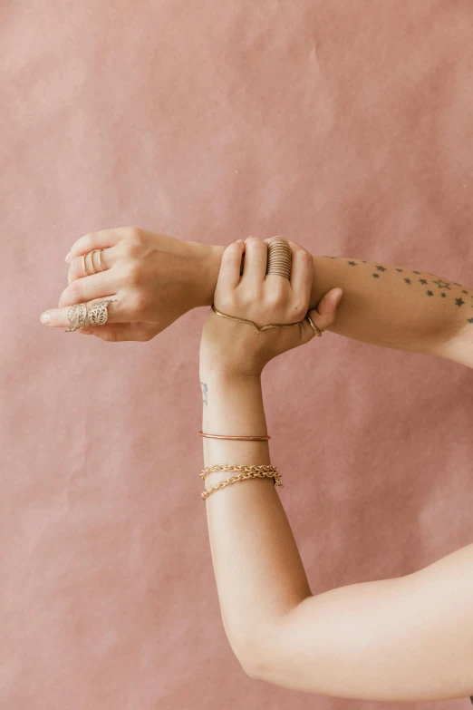 a woman with a tattoo on her arm, wearing gold jewellery, minimal pink palette, holding each other hands