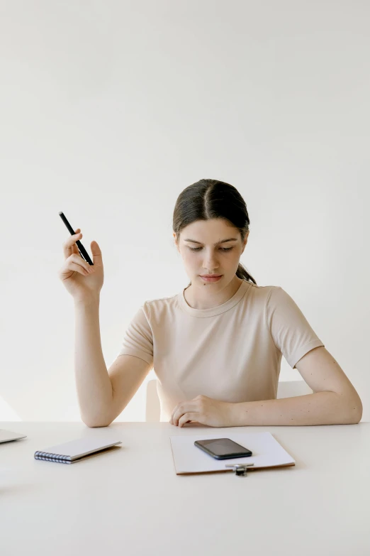 a woman sitting at a table with a cell phone in her hand, trending on pexels, holding pencil, thinking pose, wearing a light shirt, but minimalist