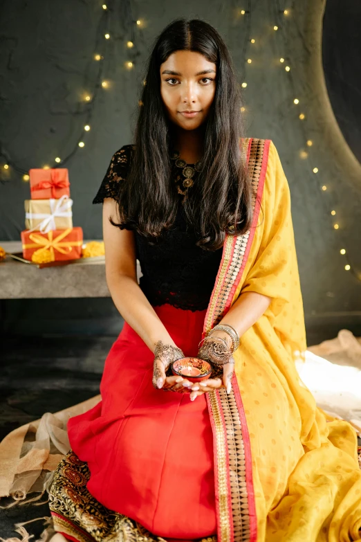 a woman in a red and yellow sari sitting on a rug, lit with candles, flatlay, wearing festive clothing, with a red skirt